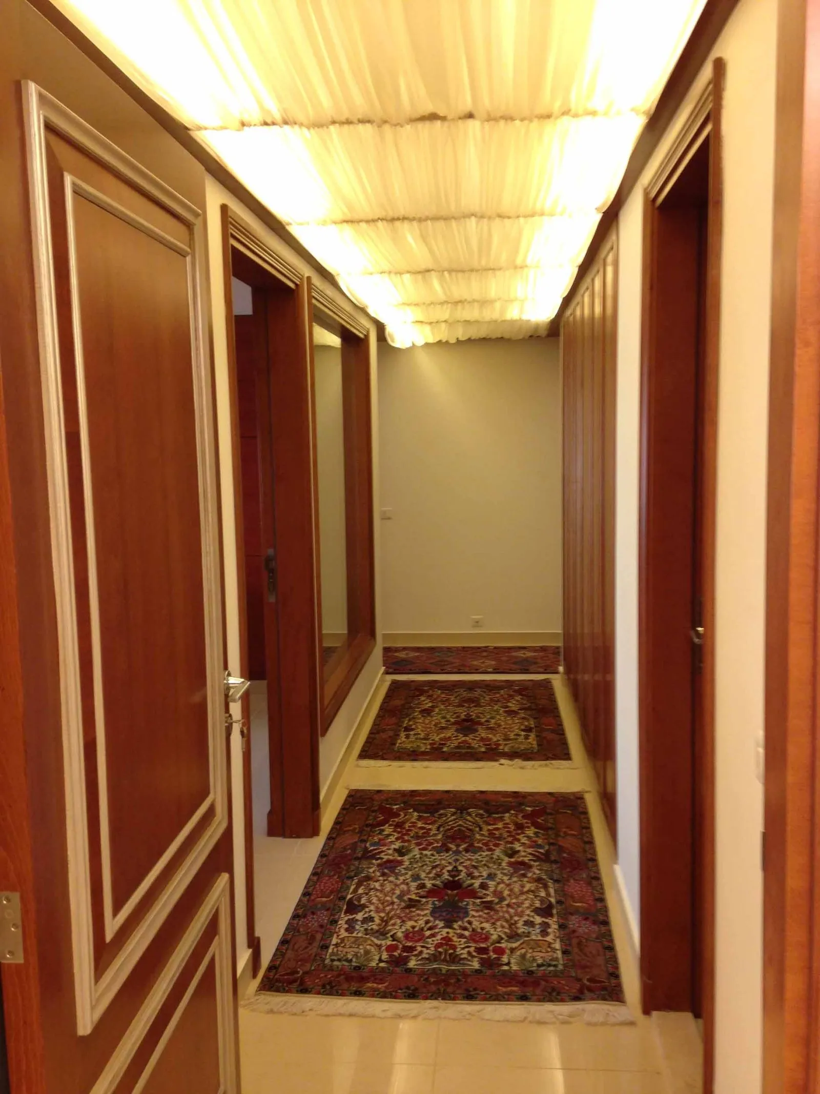 Long residential corridor with dark timber doors either side, a full-length mirror, patterned runner rugs and a draped fabric ceiling