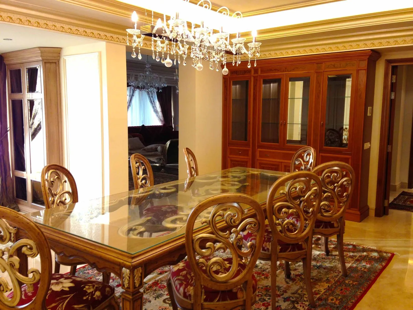 Dining room with a glass-topped carved timber table, upholstered scroll-back chairs, a crystal chandelier and mirrored display cabinets